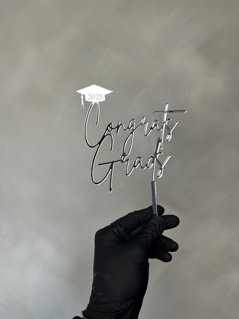 May include: A silver-colored cake topper with the words "Congrats Grads" in a script font. The year "2025" is above the words, inside a graduation cap. The topper is held by a gloved hand.