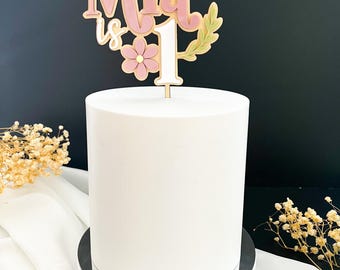 May include: A pink and white cake topper with the name "Mia is 1" and a butterfly, flowers, and leaves.
