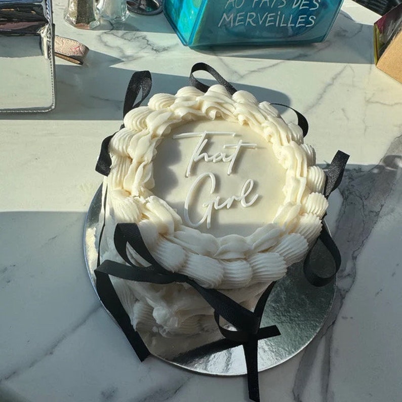 Peut inclure: Un g&acirc;teau blanc d&eacute;cor&eacute; de rubans noirs. Le g&acirc;teau est glac&eacute; de gla&ccedil;age blanc et porte l'inscription "That Girl" en gla&ccedil;age blanc.