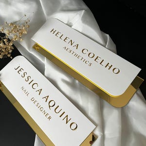 Pu&ograve; includere: Due cartelli rettangolari bianchi con bordatura dorata. Il cartello superiore recita "Helena Coelho Aesthetics" e il cartello inferiore recita "Jessica Aquino Nail Designer".
