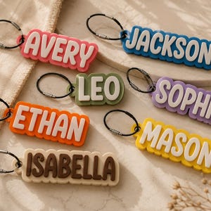 May include: A collection of rectangular keychains featuring raised white lettering on colorful backgrounds. The names Avery, Jackson, Leo, Sophia, Ethan, Mason, and Isabella are displayed in a bold, sans-serif font, each attached to a black metal ring.