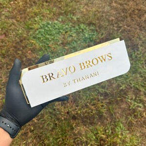 Pu&ograve; includere: Un cartello bianco con bordo dorato e il testo "BRAVO BROWS BY THANANI" in lettere dorate.