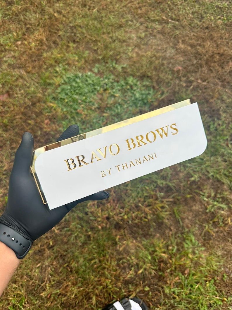 Pu&ograve; includere: Un cartello bianco con bordo dorato e il testo "BRAVO BROWS BY THANANI" in lettere dorate.