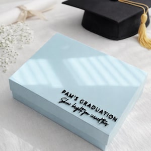 Op de afbeelding: Een lichtblauwe rechthoekige geschenkdoos met zwarte verhoogde letters met de tekst PAM'S GRADUATION en Shine bright, you earned this. De doos staat bij een zwarte afstudeerhoed met een gele kwast en een opgerold diploma.