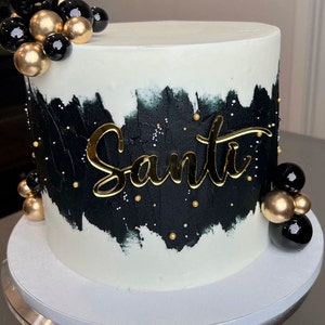 May include: A round cake with white frosting and a black and white painted design. The cake is decorated with gold and black spheres and the name "Santi" in gold script.