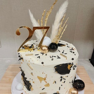 以下が含まれることがあります： 黒と金のアクセントで飾られた白いケーキ。スプリンクル、金箔、黒と金の風船などがあります。ケーキの上には金の数字27と白いパンパスグラスが飾られています。