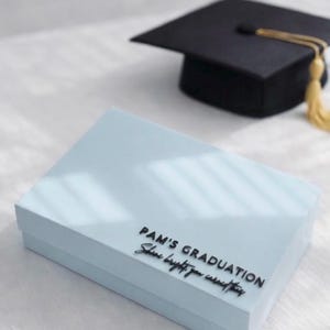 May include: A light blue rectangular gift box with black lettering that reads PAM'S GRADUATION Shine bright you earned this, positioned near a black graduation cap with a gold tassel. The box has a smooth finish and a clean, simple design.