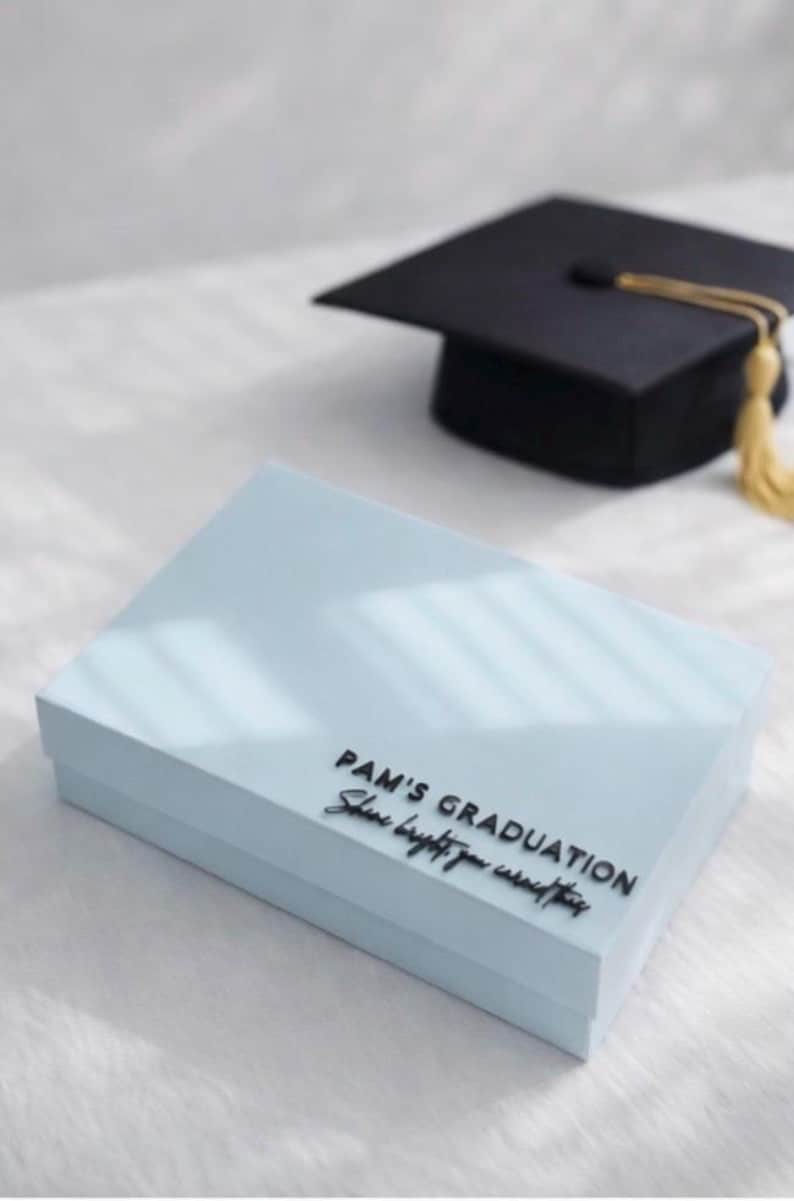 May include: A light blue rectangular gift box with black lettering that reads PAM'S GRADUATION Shine bright you earned this, positioned near a black graduation cap with a gold tassel. The box has a smooth finish and a clean, simple design.