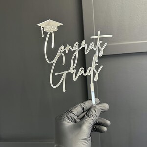 May include: A clear acrylic cake topper with the words "Congrats Grads" in a cursive font. Above the text is a graduation cap with the year "2025". The topper is held by a person wearing a black glove.