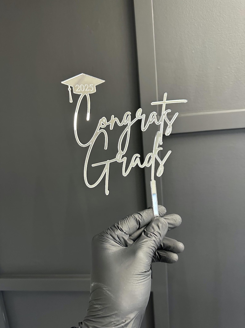 May include: A clear acrylic cake topper with the words "Congrats Grads" in a cursive font. Above the text is a graduation cap with the year "2025". The topper is held by a person wearing a black glove.