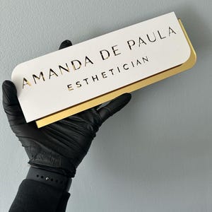 Pu&ograve; includere: Un cartello bianco con bordo dorato, con il testo "Amanda de Paula Estetista".