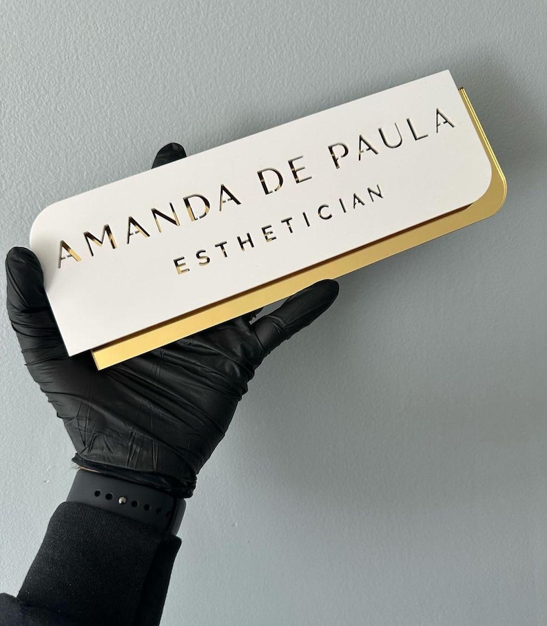 Pu&ograve; includere: Un cartello bianco con bordo dorato, con il testo "Amanda de Paula Estetista".