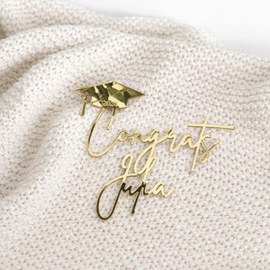 May include: Gold-colored graduation cake toppers. One topper features a graduation cap with "2024" inscribed. The other toppers spell out "Congrats Julia". The toppers are placed on a white, textured surface.