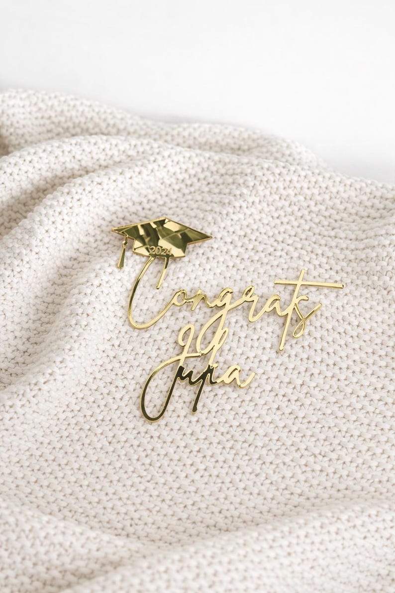May include: Gold-colored graduation cake toppers. One topper features a graduation cap with "2024" inscribed. The other toppers spell out "Congrats Julia". The toppers are placed on a white, textured surface.