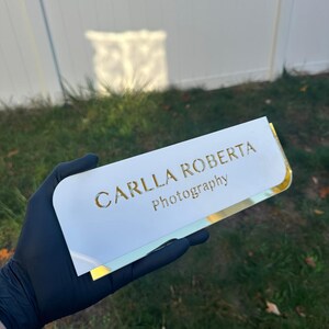 Pu&ograve; includere: Porta biglietti da visita in acrilico bianco con bordi dorati e il testo "CARLLA ROBERTA Photography" in lettere dorate.
