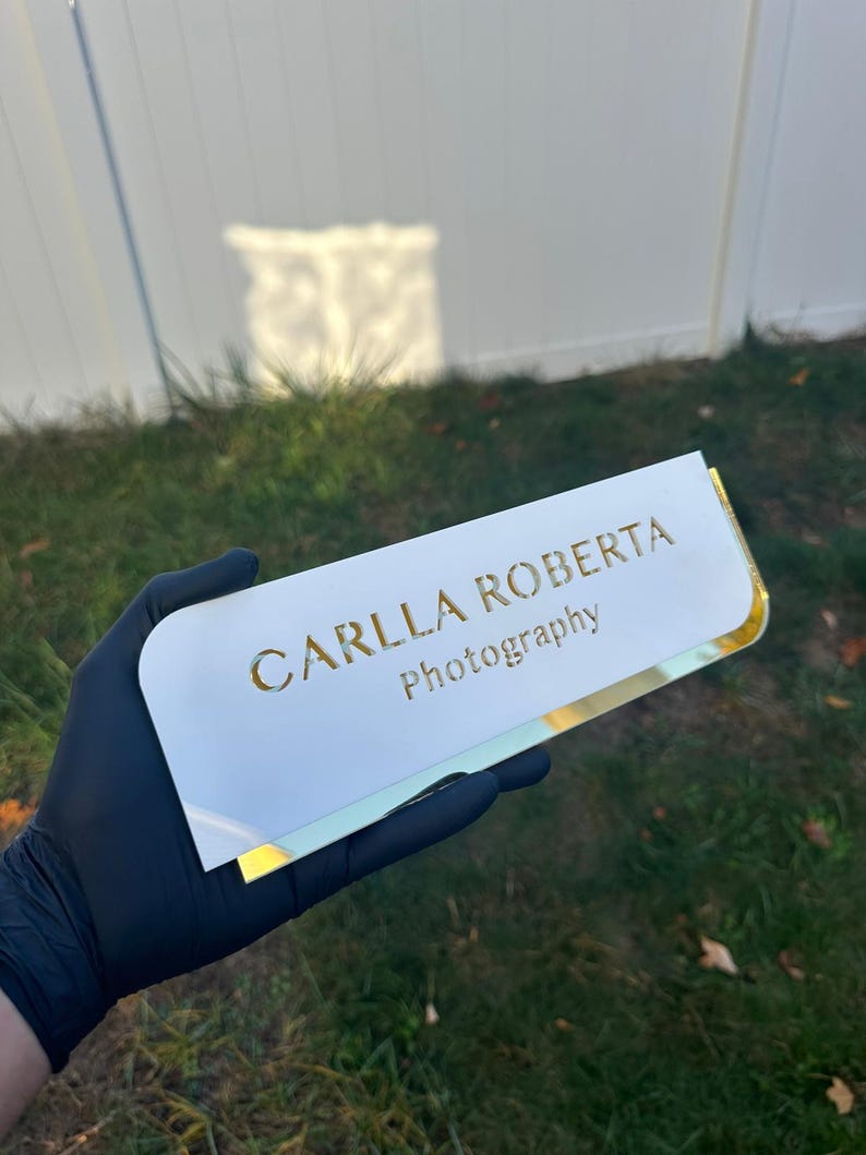 Pu&ograve; includere: Porta biglietti da visita in acrilico bianco con bordi dorati e il testo "CARLLA ROBERTA Photography" in lettere dorate.