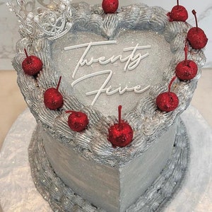 May include: A silver heart-shaped cake decorated with red cherries and the words "Twenty Five" in white icing. A silver crown sits on top of the cake.