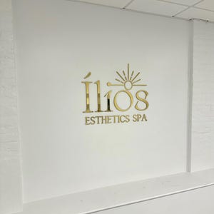 May include: Gold logo on a white wall that reads "&Iacute;lios Esthetics Spa" with a stylized sun graphic above the text.