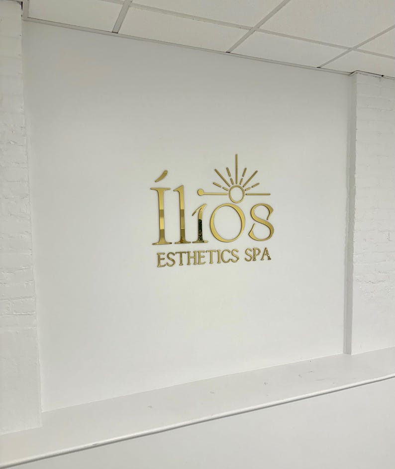 May include: Gold logo on a white wall that reads "&Iacute;lios Esthetics Spa" with a stylized sun graphic above the text.