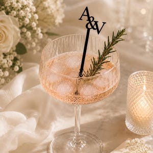 May include: A clear, ribbed glass filled with a pale pink liquid and ice cubes. A black stirring stick with the letters A&V at the top is placed inside the glass, accompanied by a sprig of green rosemary as a garnish for the drink.