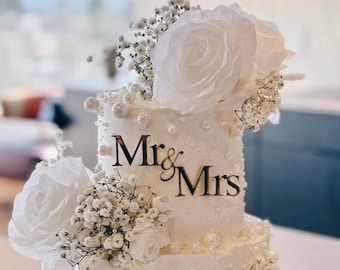 May include: A tiered white wedding cake decorated with white roses, baby's breath, and pearl accents. The words "Mr. & Mrs." are displayed in elegant black script on the front of the cake. A beautiful centerpiece for a wedding celebration.