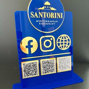 May include: Blue acrylic sign with gold lettering that reads "Santorini Mediterranean Restaurant". The sign also features gold icons for Facebook, Instagram, and a globe, as well as three QR codes.