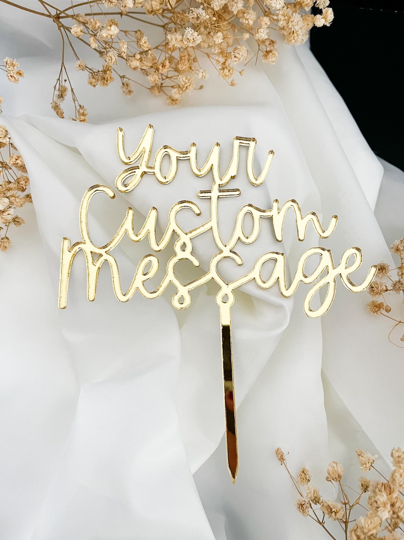 K&ouml;nnte beinhalten: Ein goldfarbener Tortenstecker aus Acryl mit dem Text Your Custom Message in einer kursiven Schriftart. Der Tortenstecker verf&uuml;gt &uuml;ber einen einzelnen vertikalen Stab in der unteren Mitte zum Einstecken in eine Torte und hat eine glatte, reflektierende Oberfl&auml;che.