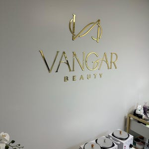 May include: Gold lettering on a white wall reads "VANGAR BEAUTY". The logo is a stylized "V" with a curved line through it.  A white wax warmer with three pots is on a table in the foreground.