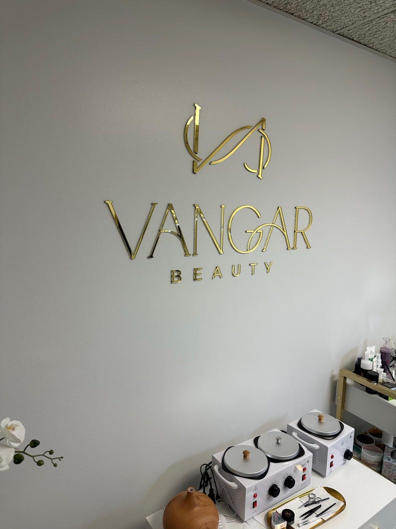 May include: Gold lettering on a white wall reads "VANGAR BEAUTY". The logo is a stylized "V" with a curved line through it.  A white wax warmer with three pots is on a table in the foreground.