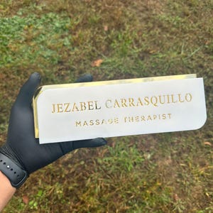 Pu&ograve; includere: Un cartello bianco con lettere dorate che recita "Jezabel Carrasquillo Massage Therapist".