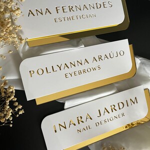 Pu&ograve; includere: Tre cartelli in acrilico bianco con bordo dorato e lettere dorate. Il cartello superiore recita "ANA FERNANDES ESTHETICIAN", il cartello intermedio recita "POLLYANNA ARAUJO EYEBROWS" e il cartello inferiore recita "INARA JARDIM NAIL DESIGNER".