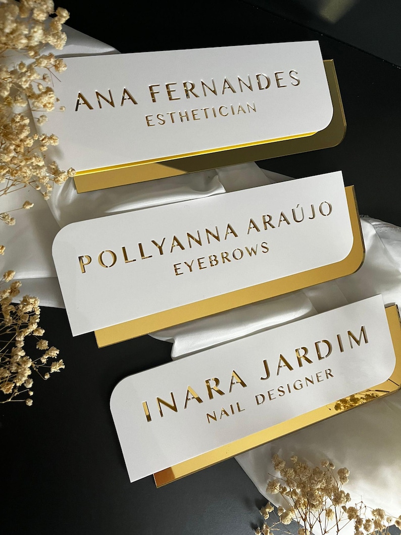 Pu&ograve; includere: Tre cartelli in acrilico bianco con bordo dorato e lettere dorate. Il cartello superiore recita "ANA FERNANDES ESTHETICIAN", il cartello intermedio recita "POLLYANNA ARAUJO EYEBROWS" e il cartello inferiore recita "INARA JARDIM NAIL DESIGNER".