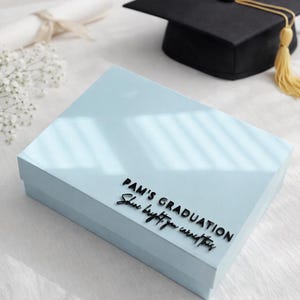 May include: A light blue rectangular gift box featuring black raised lettering that reads PAM'S GRADUATION and Shine bright, you earned this. The box is positioned near a black graduation cap with a yellow tassel and a rolled diploma.