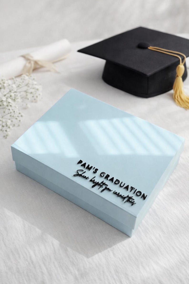 May include: A light blue rectangular gift box featuring black raised lettering that reads PAM'S GRADUATION and Shine bright, you earned this. The box is positioned near a black graduation cap with a yellow tassel and a rolled diploma.