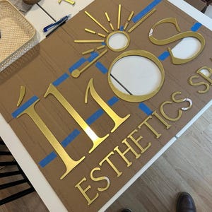 May include: Gold metal letters spelling "110" and "ESTHETICS SPA" on a brown cardboard background. Blue painter's tape is used to secure the letters to the cardboard.