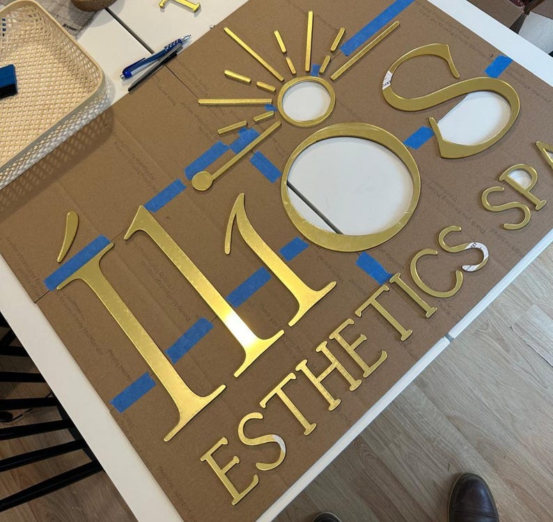 May include: Gold metal letters spelling "110" and "ESTHETICS SPA" on a brown cardboard background. Blue painter's tape is used to secure the letters to the cardboard.