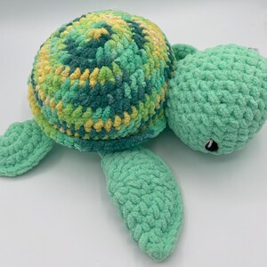 Puede incluir: Un peluche de tortuga hecho a mano con ganchillo. El caparazón es una mezcla de hilo verde, amarillo y azul, mientras que la cabeza, las aletas y el cuerpo son de color verde claro liso. La tortuga tiene un pequeño ojo negro.