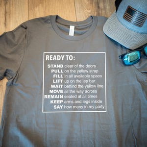 May include: Grey t-shirt with white text instructions. A blue and black hat with an American flag patch and blue sunglasses are on the shirt. The text includes instructions.
