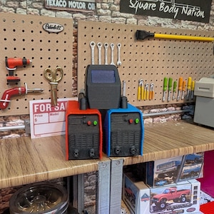 May include: A miniature workshop scene with a pegboard wall featuring tools, a Texaco sign, and a sign that reads "Square Body Nation". Two red and blue welding machines are on a wooden workbench with a tire and a model truck box.