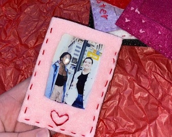 May include: A pink felt photo frame with a red stitched heart and a photo of two people standing in front of a building. The frame is surrounded by other felt pieces in various colors.