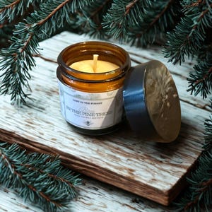 Op de afbeelding: Een bruine glazen potkaars met een deksel, met het opschrift "Lost in the Forest" en "In the Pine Trees". De kaars staat op een houten oppervlak, omringd door dennentakken. De kaars heeft gele was en een witte lont.