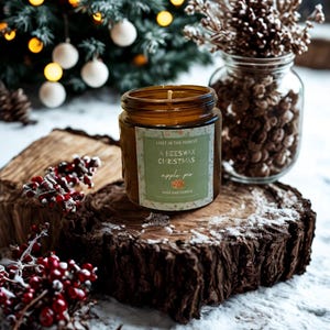 May include: A brown glass jar candle labeled "Lost in the Forest, A Beeswax Christmas, apple pie." The candle sits on a wood slice with red berries and a Christmas tree in the background. A glass jar with decorative pinecones is also visible.