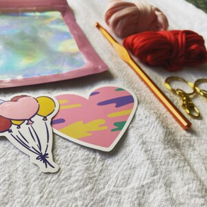 May include: A close-up of a craft project with colorful yarn, a crochet hook, and two stickers. One sticker is a heart with a pink, yellow, and purple abstract design. The other sticker is a bunch of colorful balloons.