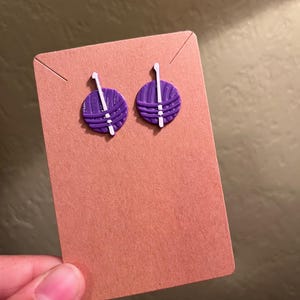 May include: A pair of purple yarn ball earrings with white knitting needles. The earrings are on a brown card.
