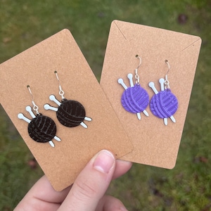 May include: Two pairs of dangle earrings featuring a yarn ball with knitting needles. One pair is purple and the other is black.