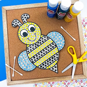 May include: A framed corkboard features a bee-shaped craft project with yellow, black, and blue dot patterns. Nearby are blue, black, and yellow paint bottles, cotton swabs, and yellow scissors, suggesting a DIY craft activity.