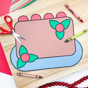 May include: A craft project featuring a paper cake with pink frosting, green holly accents, and a blue base. Red scissors and crayons in pink, brown, and green are placed around the cake on a wooden surface. Decorative paper and red beads are also visible.