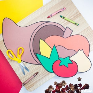 May include: A craft project for making a cornucopia out of paper. The cornucopia is made of brown, orange, and yellow paper. There are also green leaves, a red tomato, and a white onion. The project is laid out on a wooden surface.