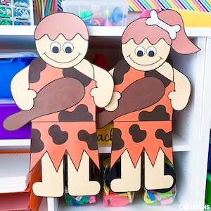 May include: Two large cardboard cutouts of cave people, one with brown hair and one with pink hair, both holding a brown club. The cutouts are standing on a white shelf with colourful containers below.