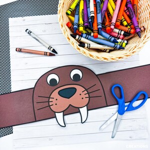 May include: A brown walrus headband craft with white tusks, black whiskers, and large eyes. A basket of crayons in various colors sits above the headband. Two crayons and blue scissors are also on the white wooden surface.